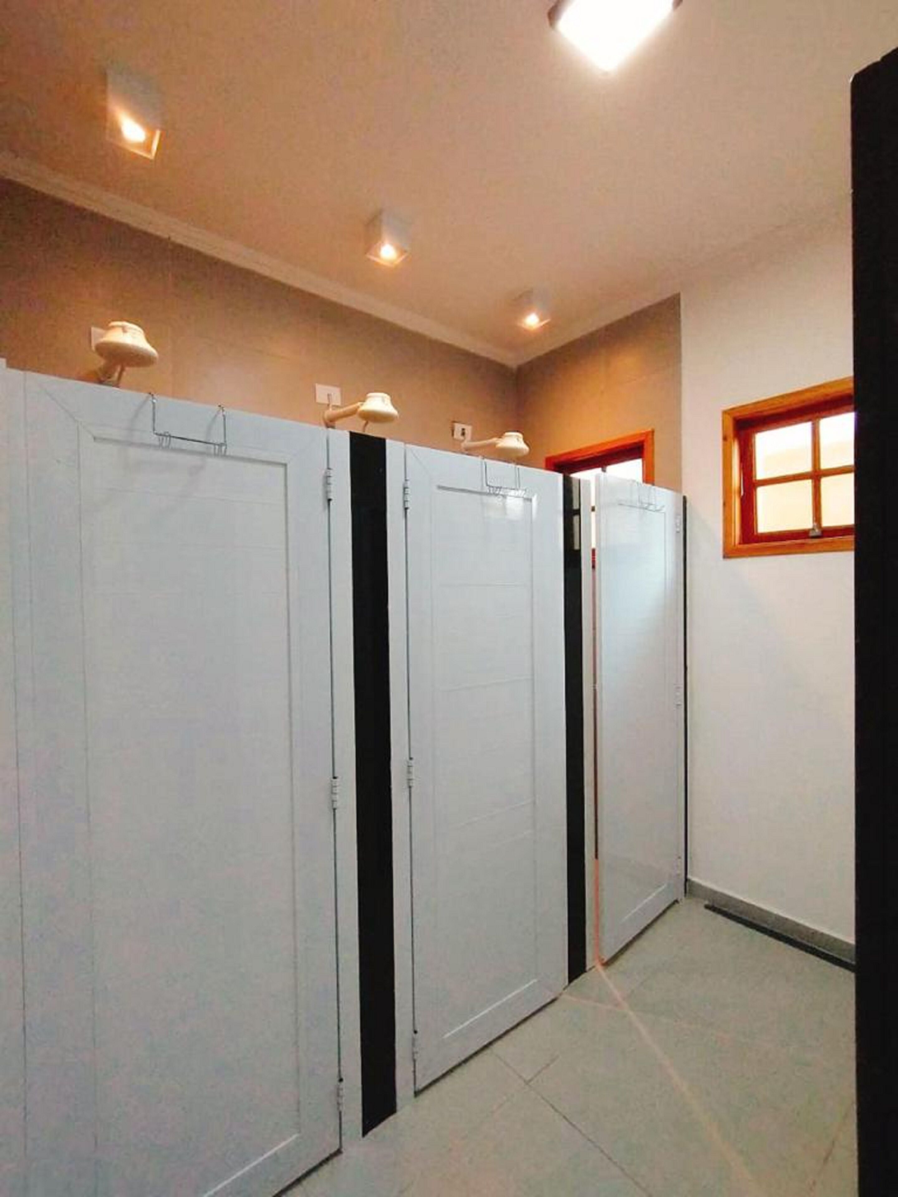 A hallway with four white doors, each with a black vertical stripe. There are ceiling lights and a window with wooden frames on the right wall.