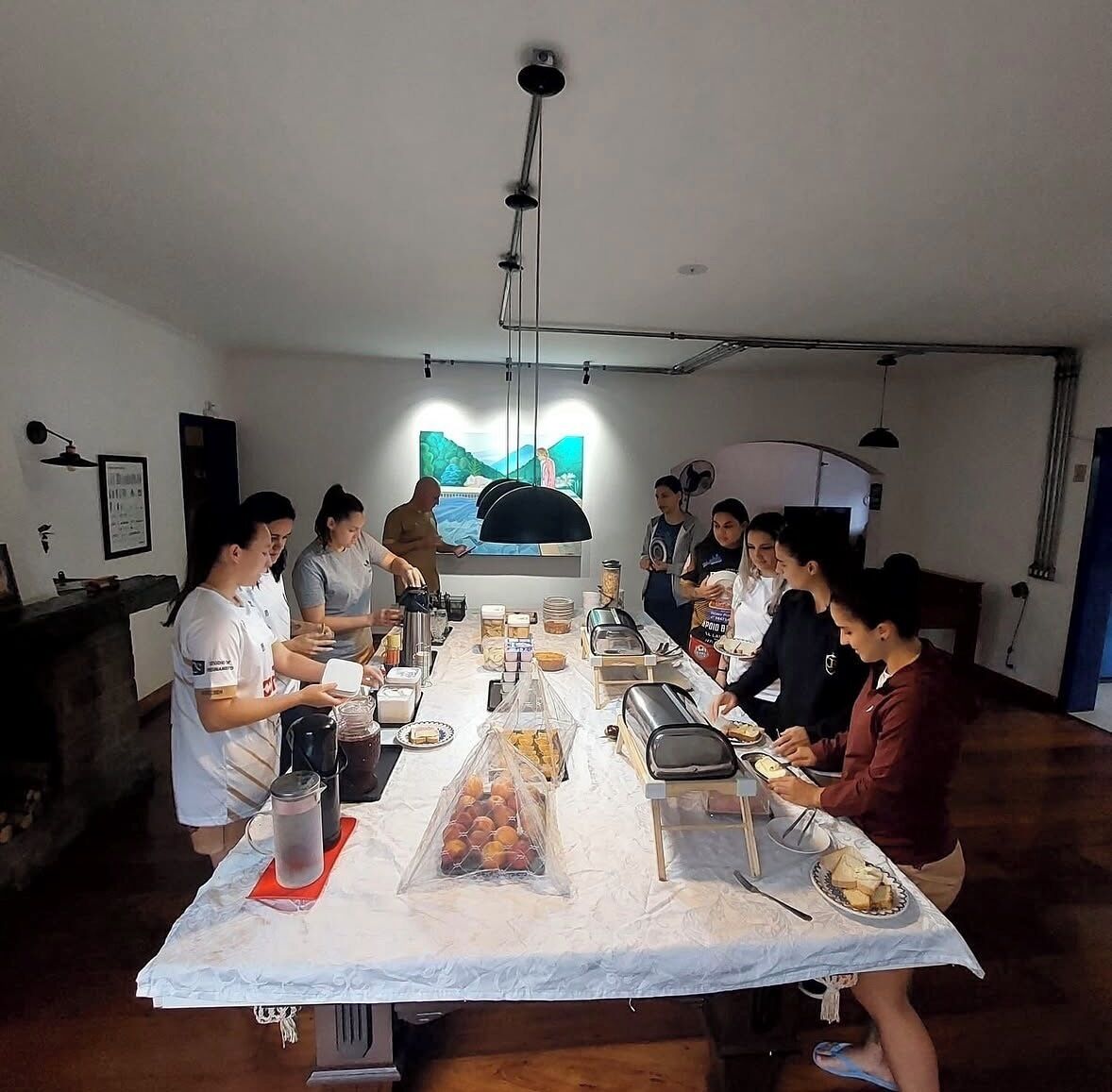 Un comedor compartido con una larga mesa, distintos alimentos y un cuadro en la pared.
