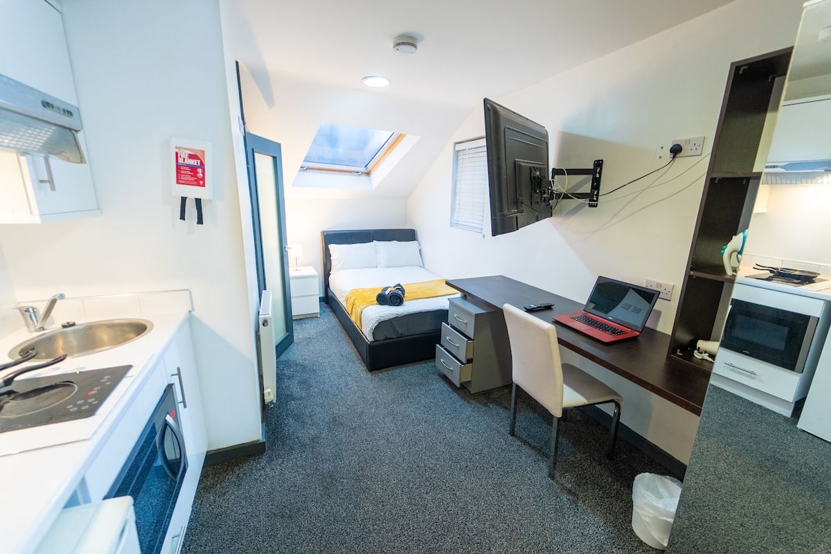 Une chambre d’hôtel compacte comprenant un coin cuisine, un lit avec une couverture jaune, un bureau avec un ordinateur portable et une petite fenêtre.