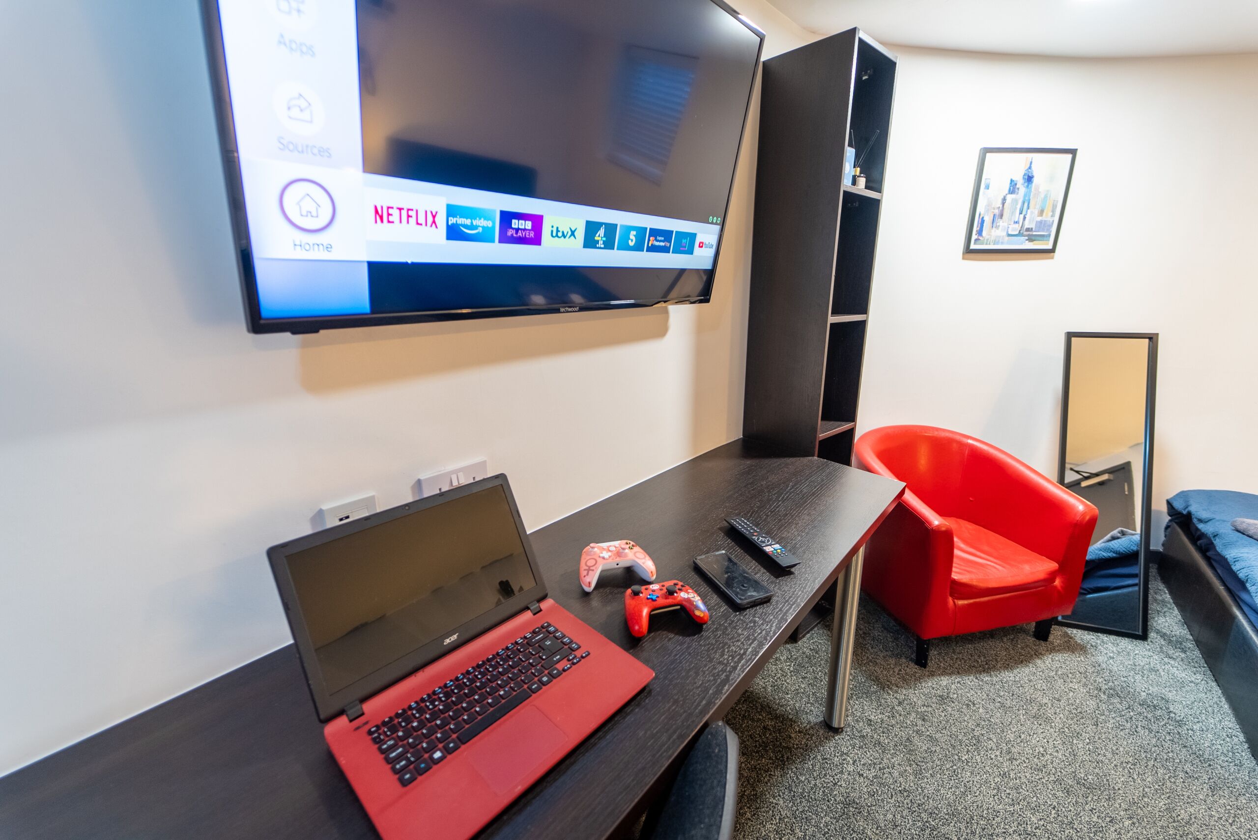 A modern room with a flat-screen TV mounted on the wall displaying Netflix and other streaming services. A laptop and gaming controller are on a desk, and a red chair is positioned near the TV.