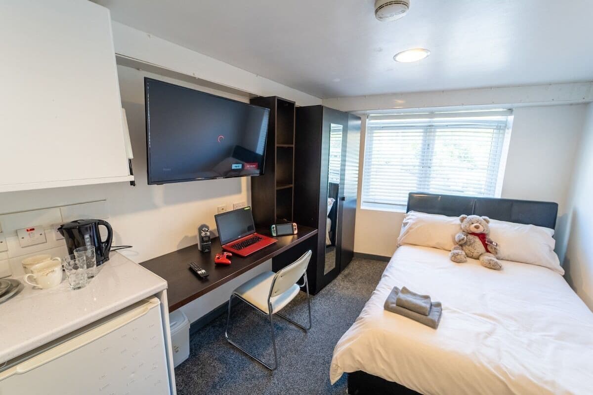 Une chambre d’hôtel compacte comprenant un lit, un bureau avec un ordinateur portable, une télévision et une fenêtre avec des stores.