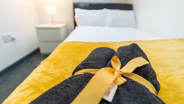 Un lit bien fait, recouvert d’une couverture jaune et agrémenté d’une serviette noire ornée d’un ruban doré.