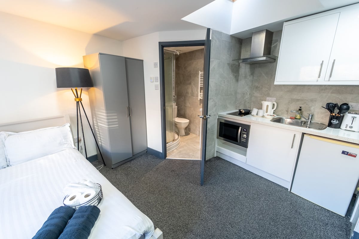 Une chambre d’hôtel de taille réduite, comprenant un lit, une lampe de chevet, une petite kitchenette et une salle de bain avec des toilettes et une douche.
