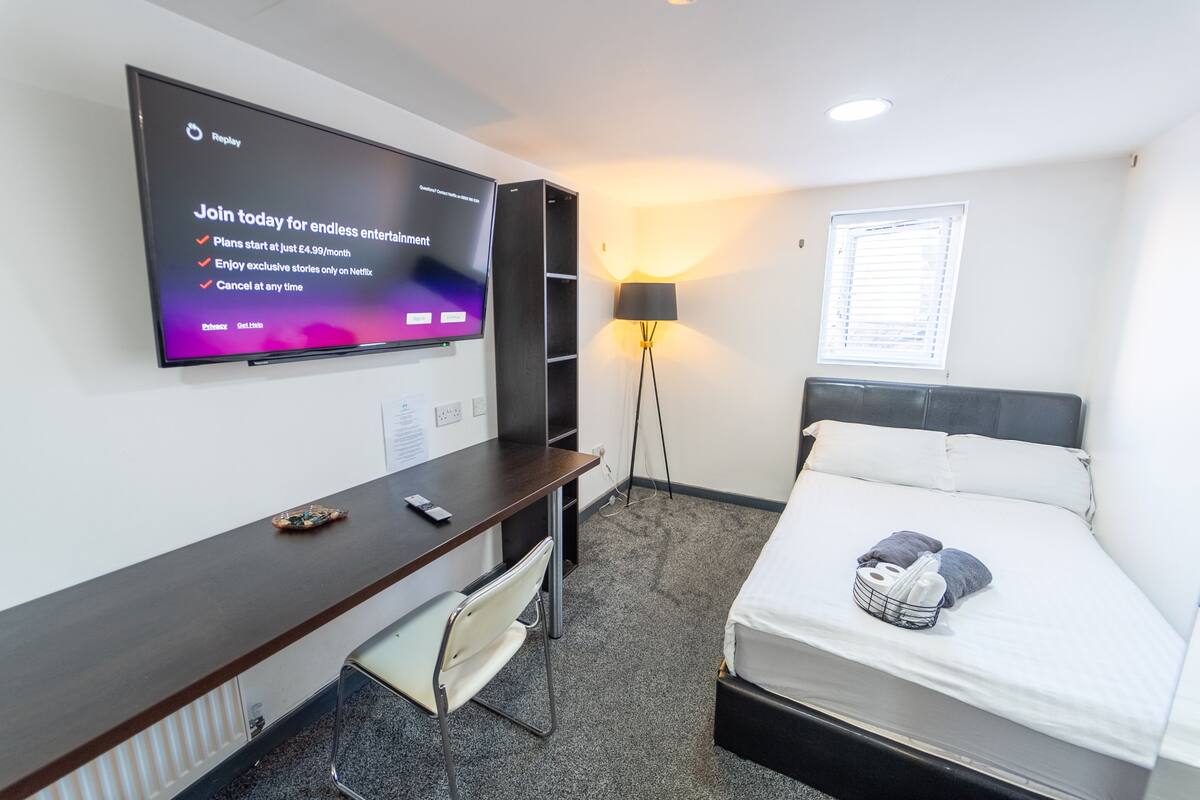 Une chambre d’hôtel avec un lit, un bureau, une chaise, un téléviseur diffusant une publicité Netflix, une lampe et une fenêtre avec des stores.