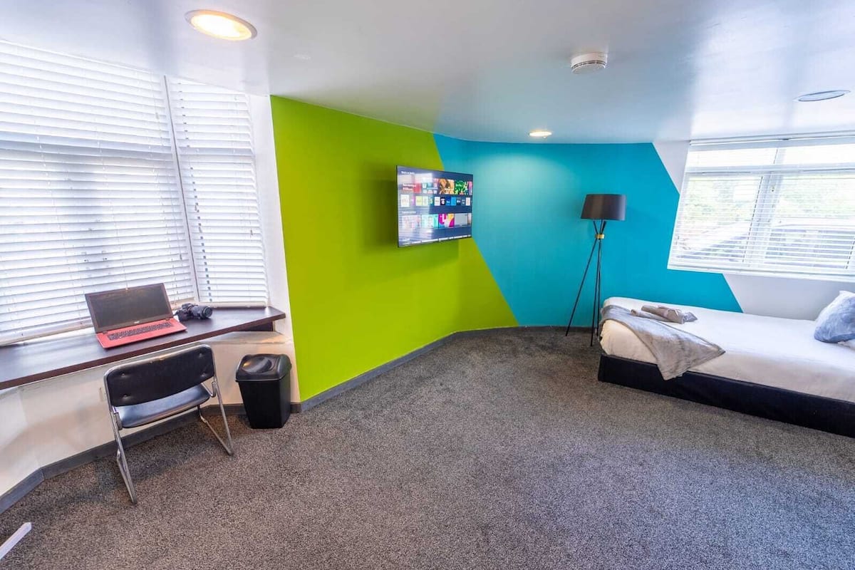 Une chambre d’hôtel avec un lit, un bureau avec un ordinateur portable, une télévision et une poubelle.