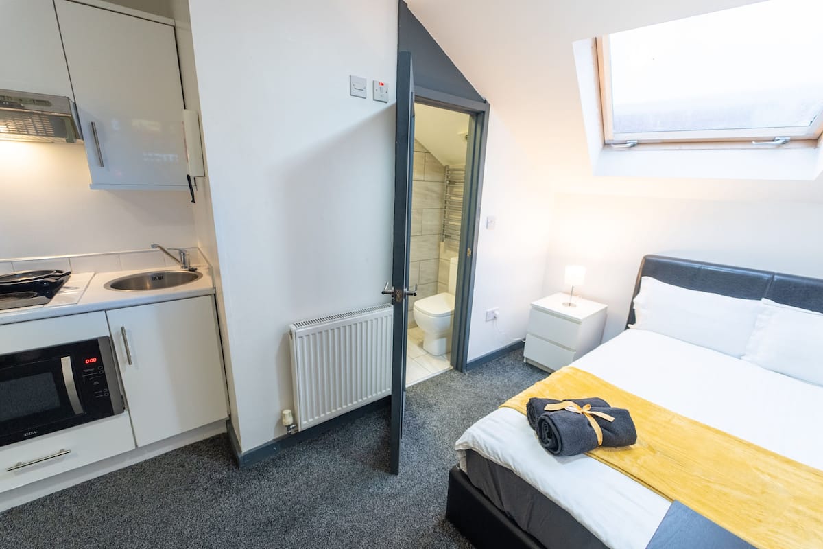 Une chambre d’hôtel avec une kitchenette, un lit doté d’une tête de lit en bois, une table de chevet et une fenêtre.