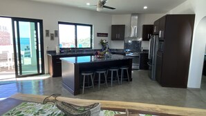 A modern kitchen with a large island, dark cabinetry, and a view of the ocean through the sliding glass doors.