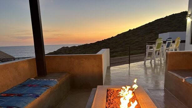 A fire pit with a view of the ocean at sunset, outdoor seating with chairs, and a concrete floor.