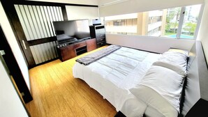 A modern bedroom with a large bed, a TV, a dresser, and a window with a view of buildings.