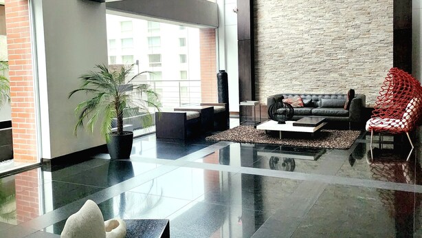 A spacious lobby with a high ceiling, a black leather sofa, a red chair, a potted plant, and a white sculpture.