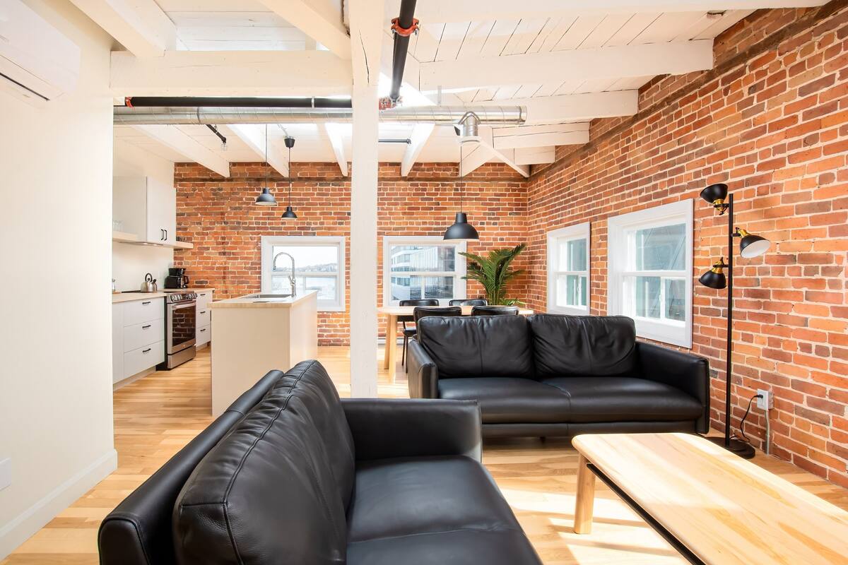 Un espace de vie moderne avec un mur en briques, des canapés en cuir noir, un banc en bois et un coin repas agrémenté d’une plante.