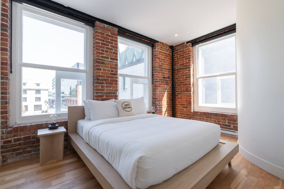Une chambre avec un lit en mezzanine, de grandes fenêtres, des murs en briques et une petite table de chevet.