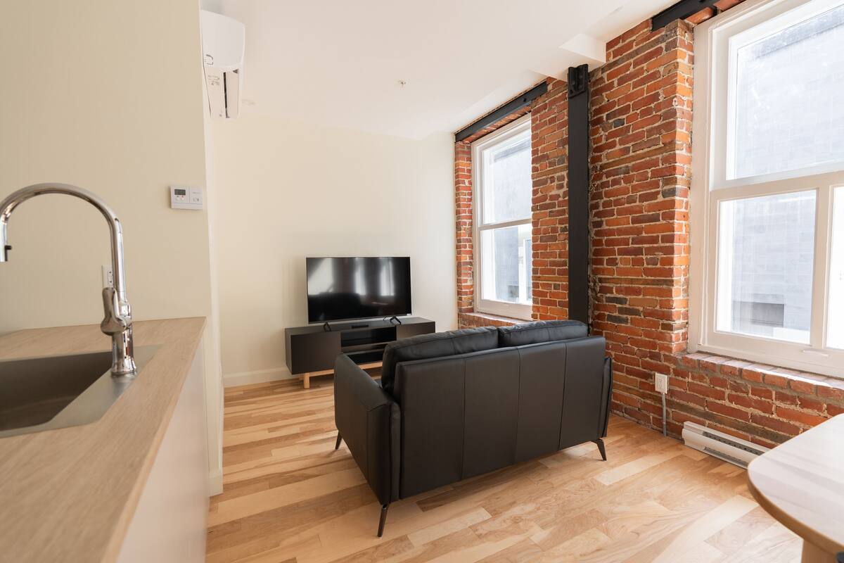 Un salon moderne avec un canapé noir, un téléviseur à écran plat, un plancher en bois et un mur en briques.