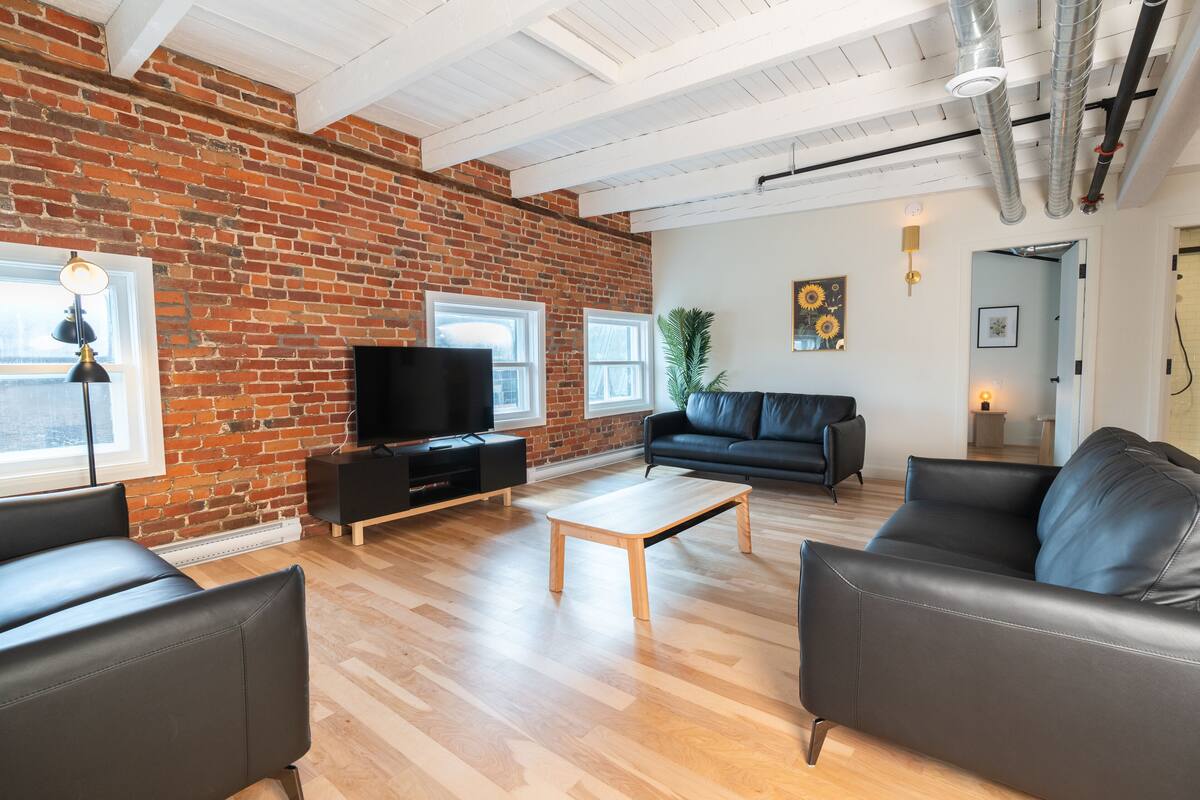 Un salon moderne avec un mur en briques, un plancher en bois, un canapé en cuir noir, une table basse et un téléviseur posé sur un meuble noir.