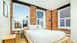 Ein Schlafzimmer mit Bett, einer hölzernen Bank und Blick auf Gebäude durch große Fenster.