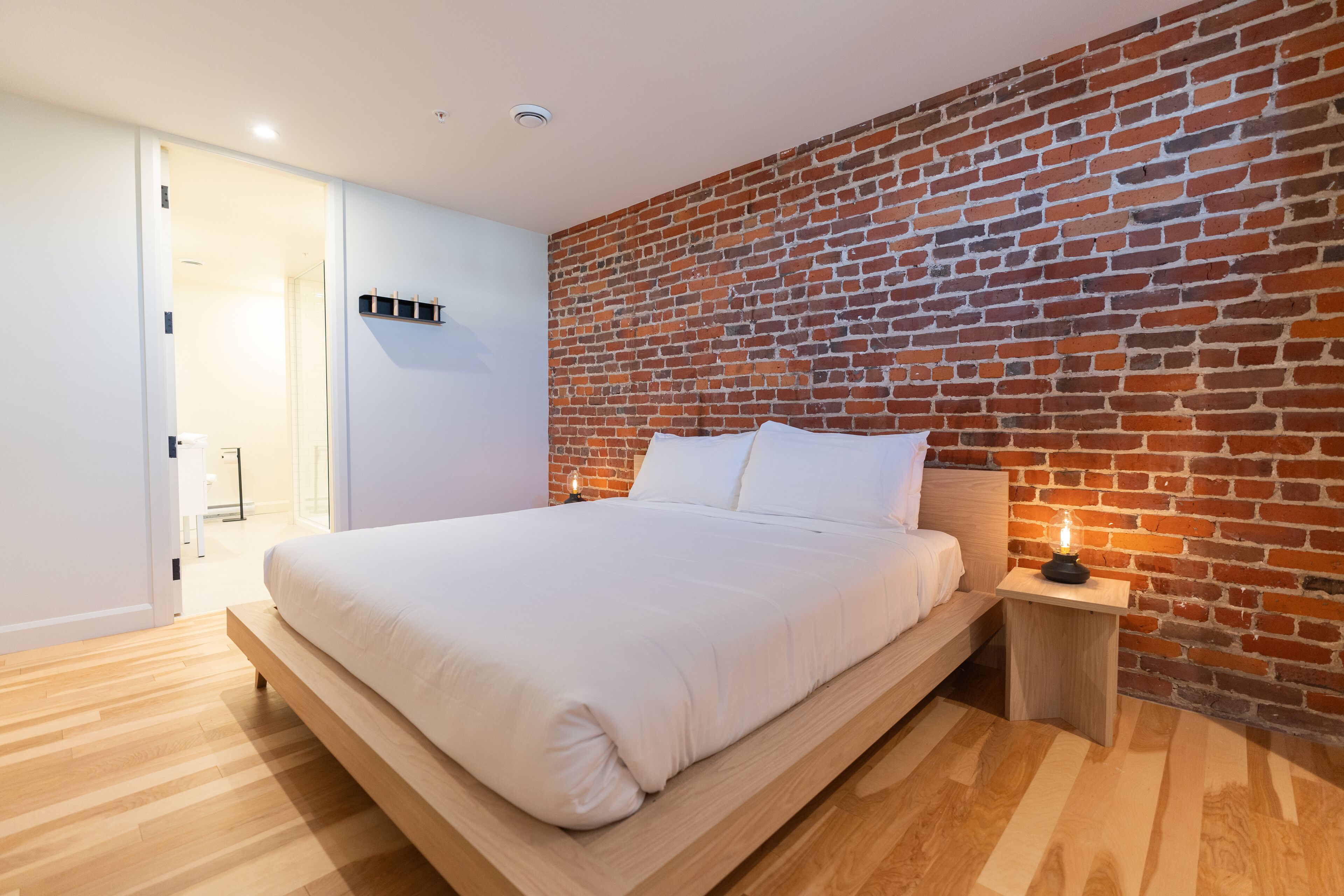 A modern bedroom with a brick accent wall, a wooden bed frame, and a bedside table with a lamp.