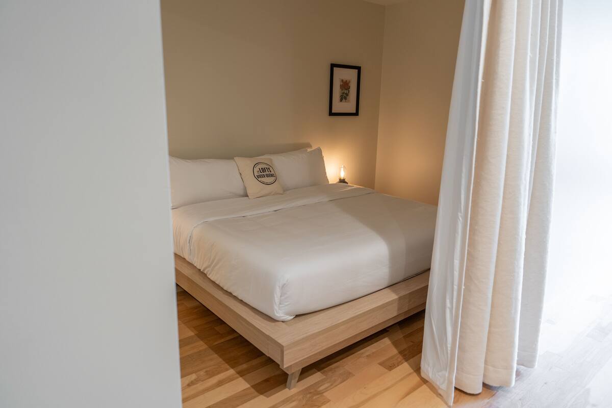 Un lit bien fait, avec du linge de lit blanc et un oreiller orné d’un logo, un tableau encadré au mur et un plancher en bois.