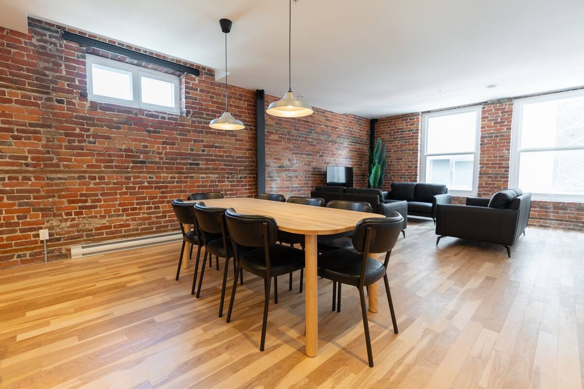 Une pièce spacieuse avec une table à manger en bois entourée de chaises, un canapé noir et un téléviseur placé contre un mur en briques.