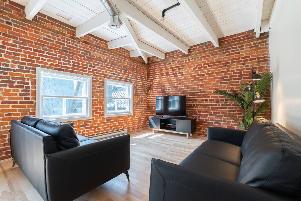 Un salon moderne avec un mur en briques, des canapés en cuir noir, un téléviseur à écran plat et une grande fenêtre.