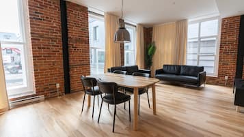 Amplia sala de estar con piso de madera, una mesa de comedor con sillas, un sofá de cuero negro y ventanas grandes con cortinas.