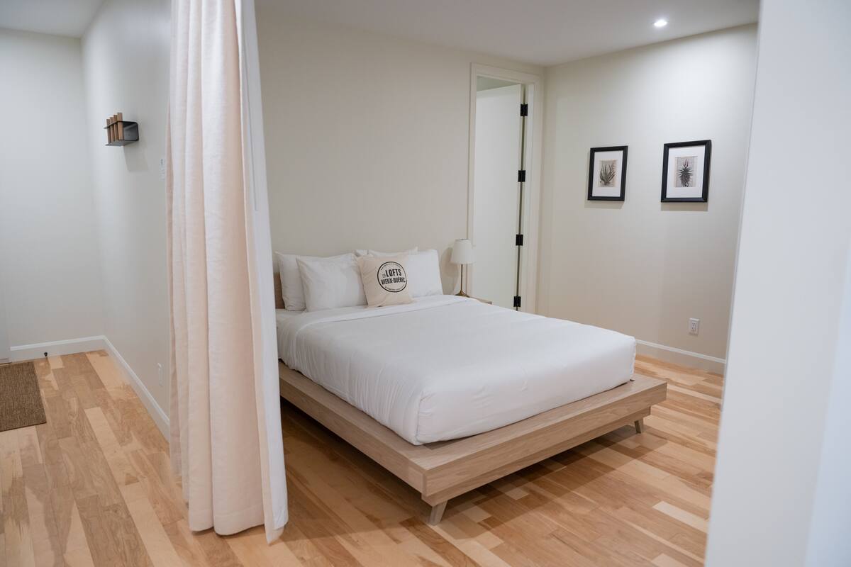 Une chambre à coucher avec un lit en bois, du linge de lit blanc, une table de chevet, une lampe et deux tableaux encadrés accrochés au mur.