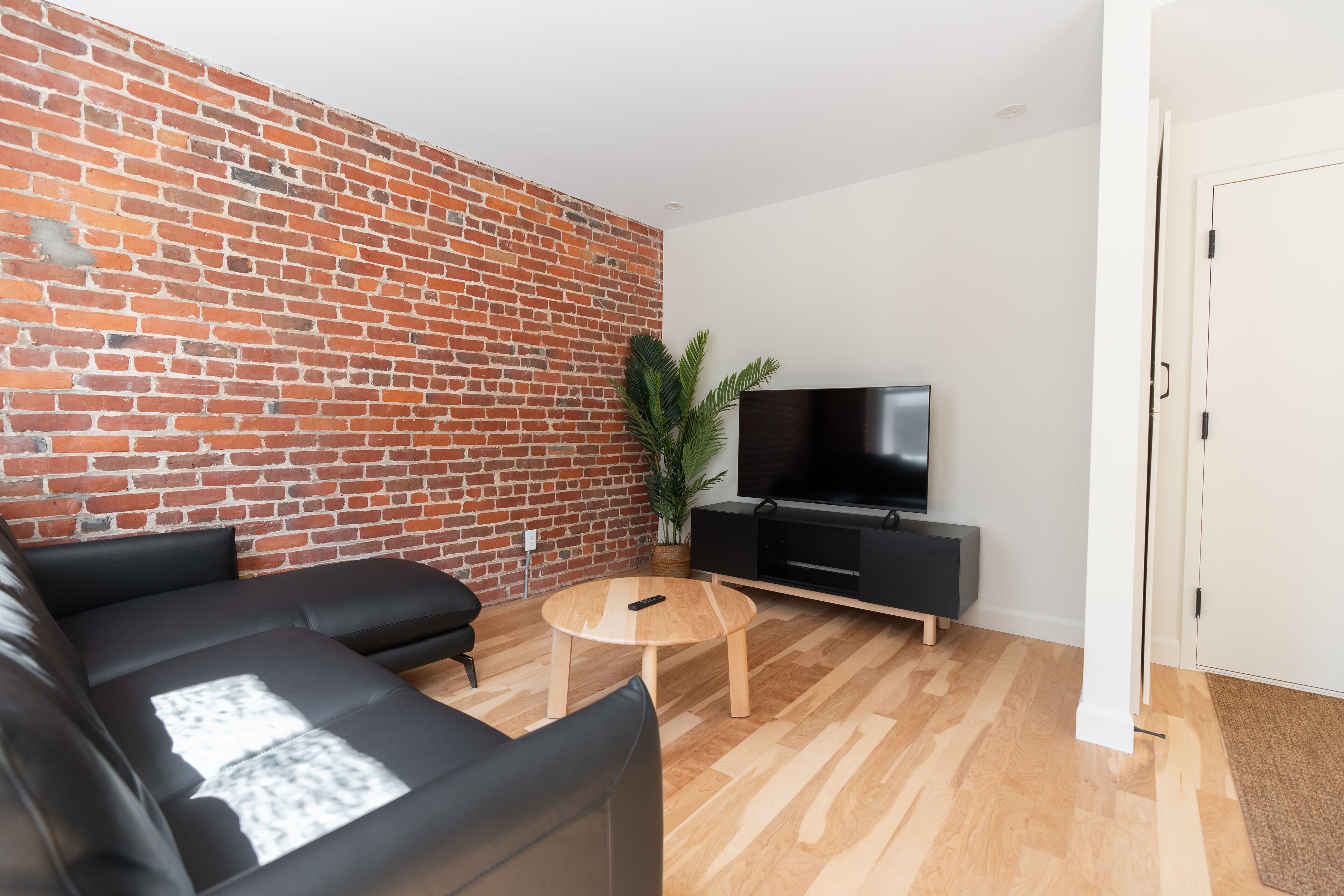 Una sala de estar moderna con pared de ladrillo, sofá de cuero negro, una mesa de centro de madera, un televisor sobre un soporte negro y una planta en maceta.