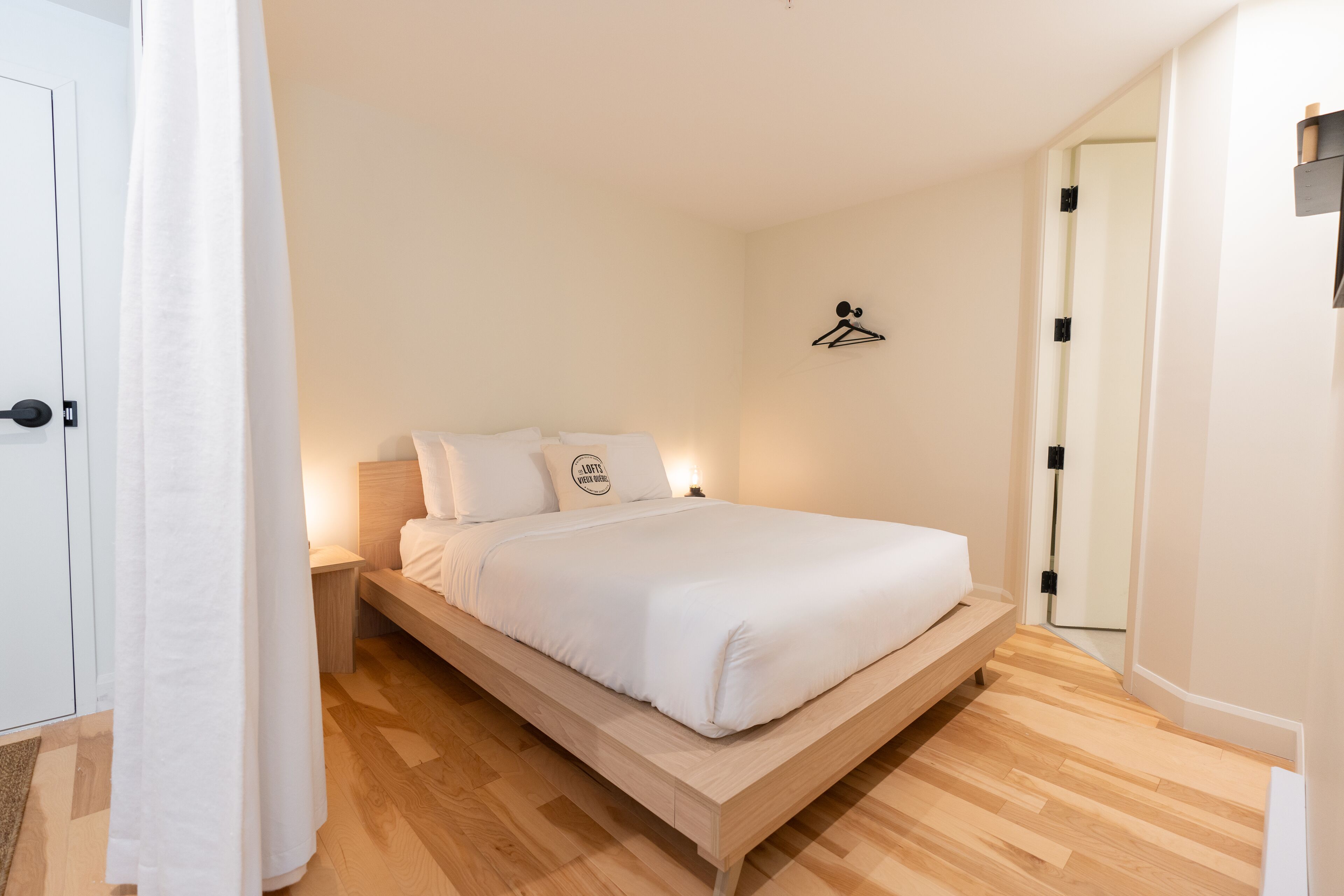 A single-bed room with a wooden frame, white bedding, a bedside table, and a wall-mounted light fixture.
