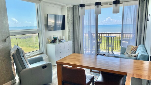Et moderne værelse med et træspisebord, to stole, en sofa, et fjernsyn og en balkon med havudsigt.