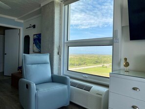 Ein Zimmer mit einem großen Fenster, einem blauen Sessel, einer weißen Kommode und einem Gemälde an der Wand.