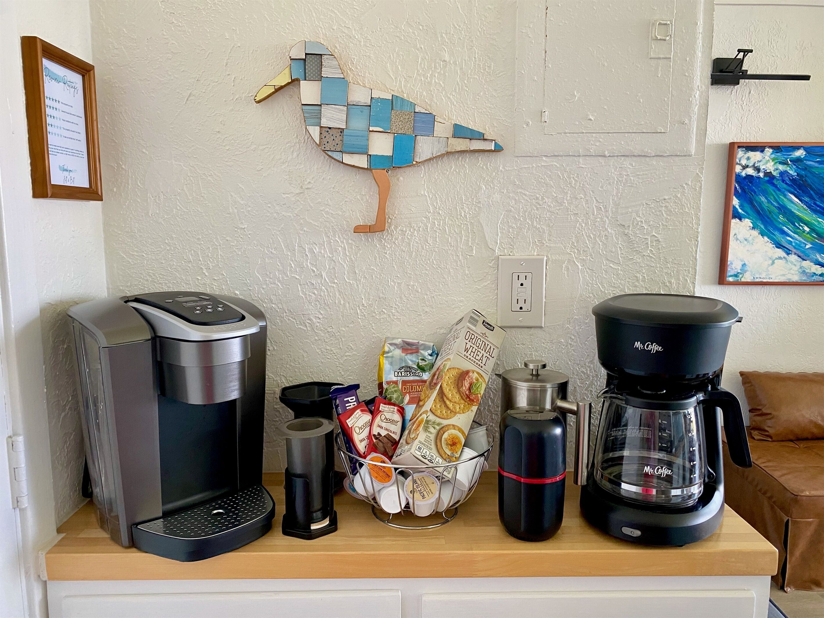 A kitchen counter with a coffee maker, a toaster, a kettle, and a coffee grinder. There is a basket of coffee pods and a box of coffee creamer.
