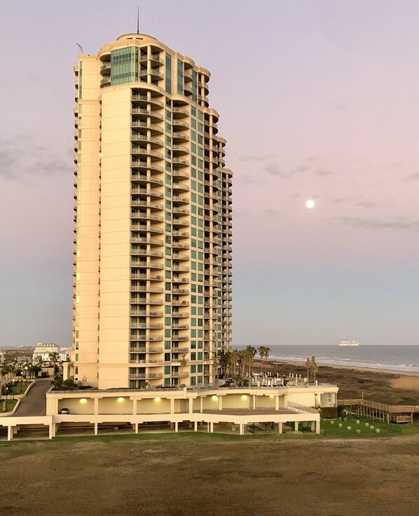 Un grand hôtel à plusieurs étages, doté de balcons, situé près d’une plage, avec un navire visible au loin.