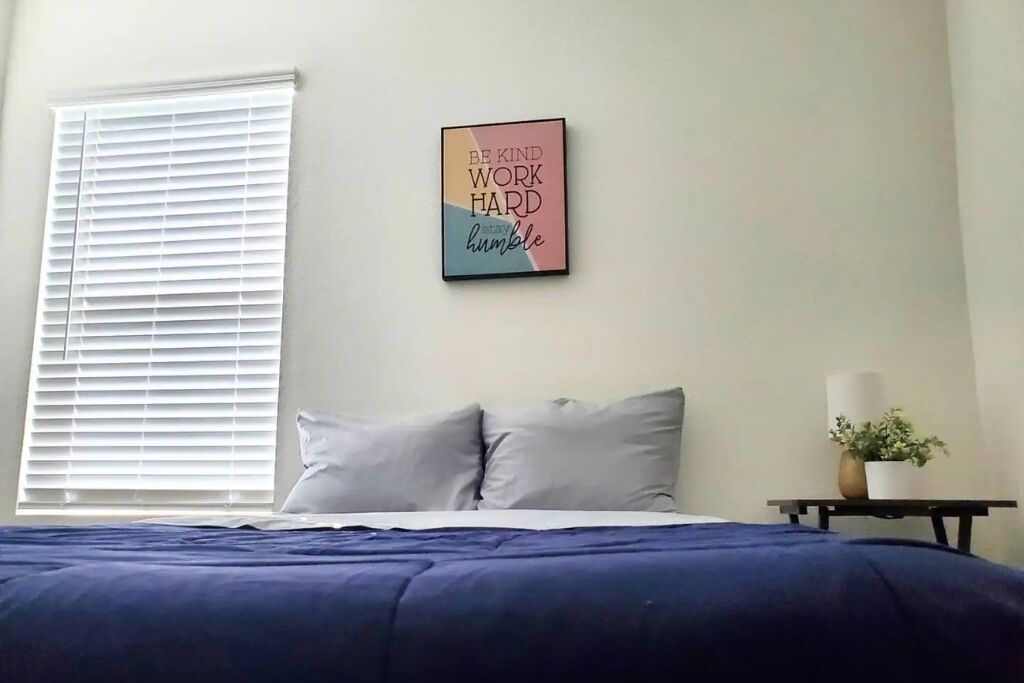 A bedroom with a bed, pillows, a bedside table with a lamp, and a wall with a motivational quote.