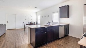A modern kitchen with a central island, dark cabinetry, and a dining area with a chair and table. - ENTIRE HOME | 3BR WFH Hub | Fast 300 Mbps Wi-Fi | Near SoCo (Austin)