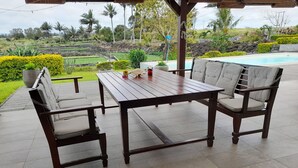An outdoor patio with a wooden table and two cushioned chairs, surrounded by a tiled floor and a view of a swimming pool and greenery. - Jags villa (Union Vale)