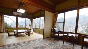 Una habitación con esteras de tatami, una mesa baja de tatami, una silla de tatami, un escritorio de madera y vistas a través de amplios ventanales.