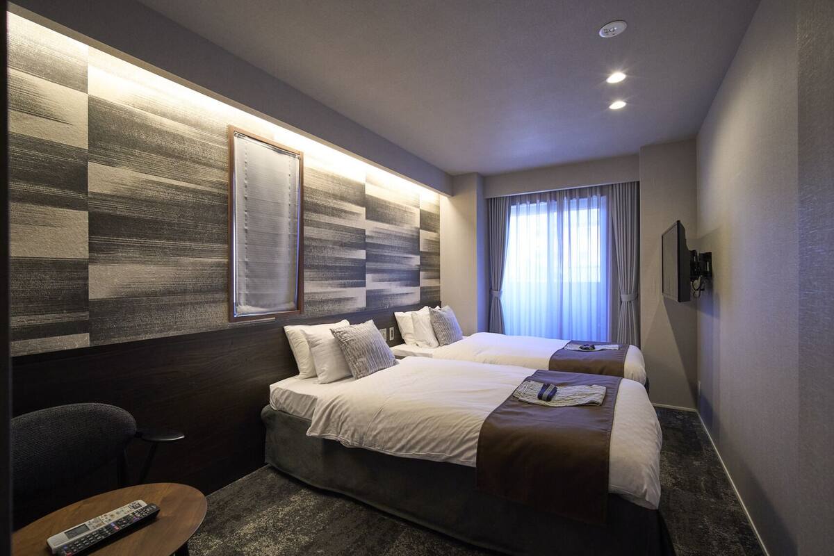 Une chambre d’hôtel avec deux lits, une télévision, une télécommande et une grande fenêtre avec des rideaux.