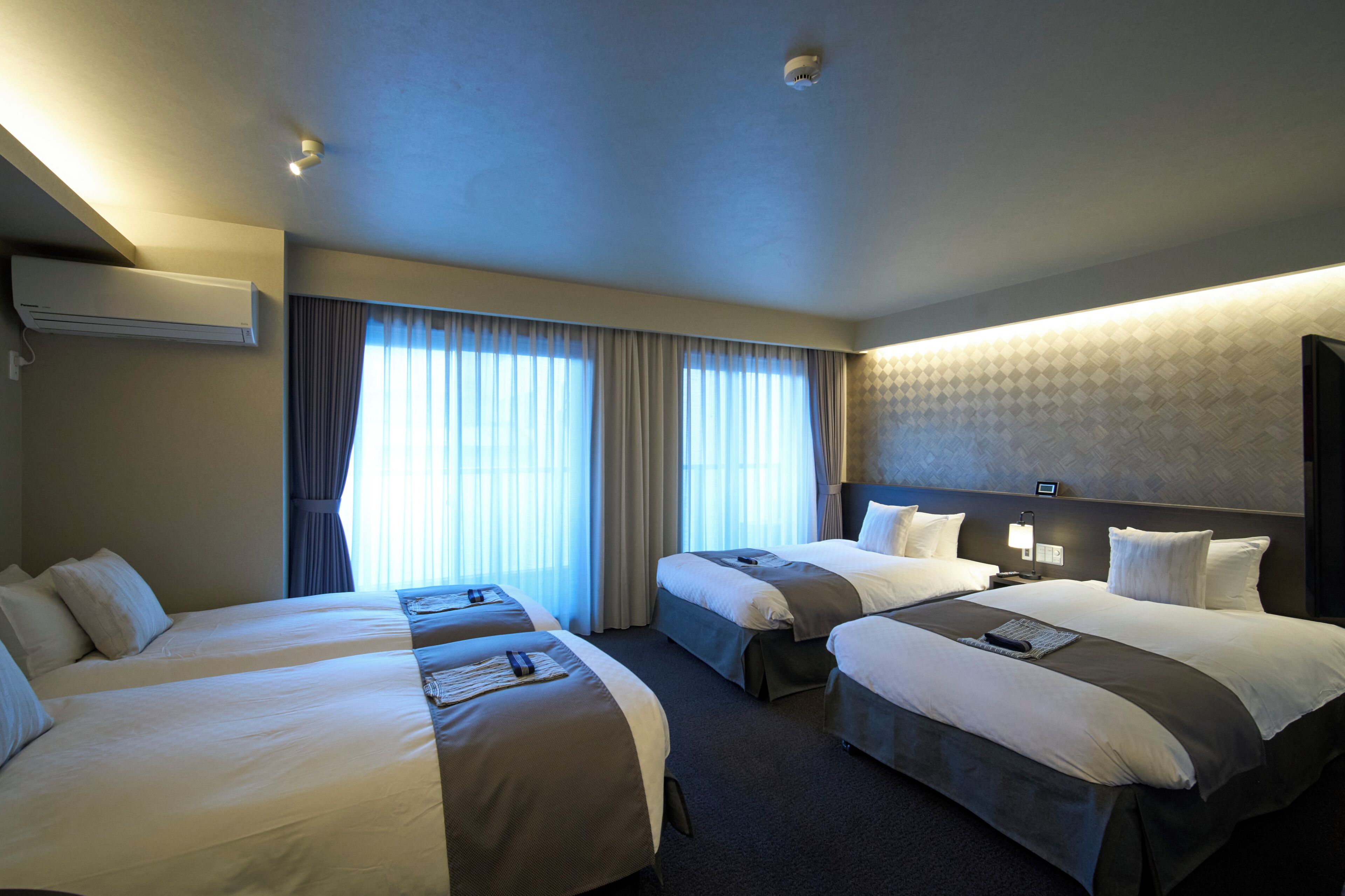 Habitación de hotel con dos camas, un ventanal grande con cortinas, unidad de aire acondicionado y cabecero con lámpara.