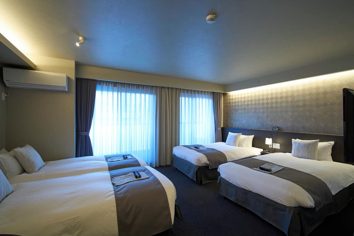 Une chambre d’hôtel avec deux lits, une grande fenêtre avec des rideaux, un appareil de climatisation et une tête de lit dotée d’un luminaire.