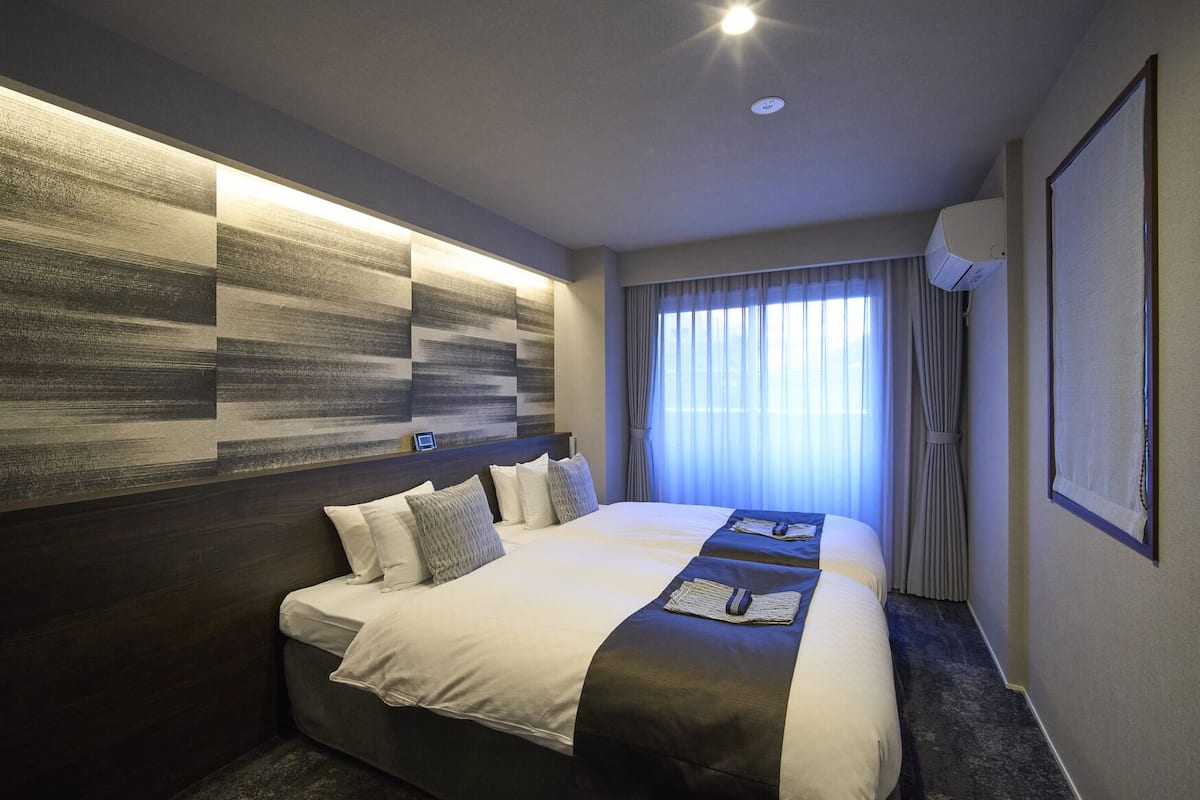 Une chambre d’hôtel avec un grand lit, une tête de lit au motif texturé, une fenêtre avec des rideaux et un climatiseur fixé au mur.