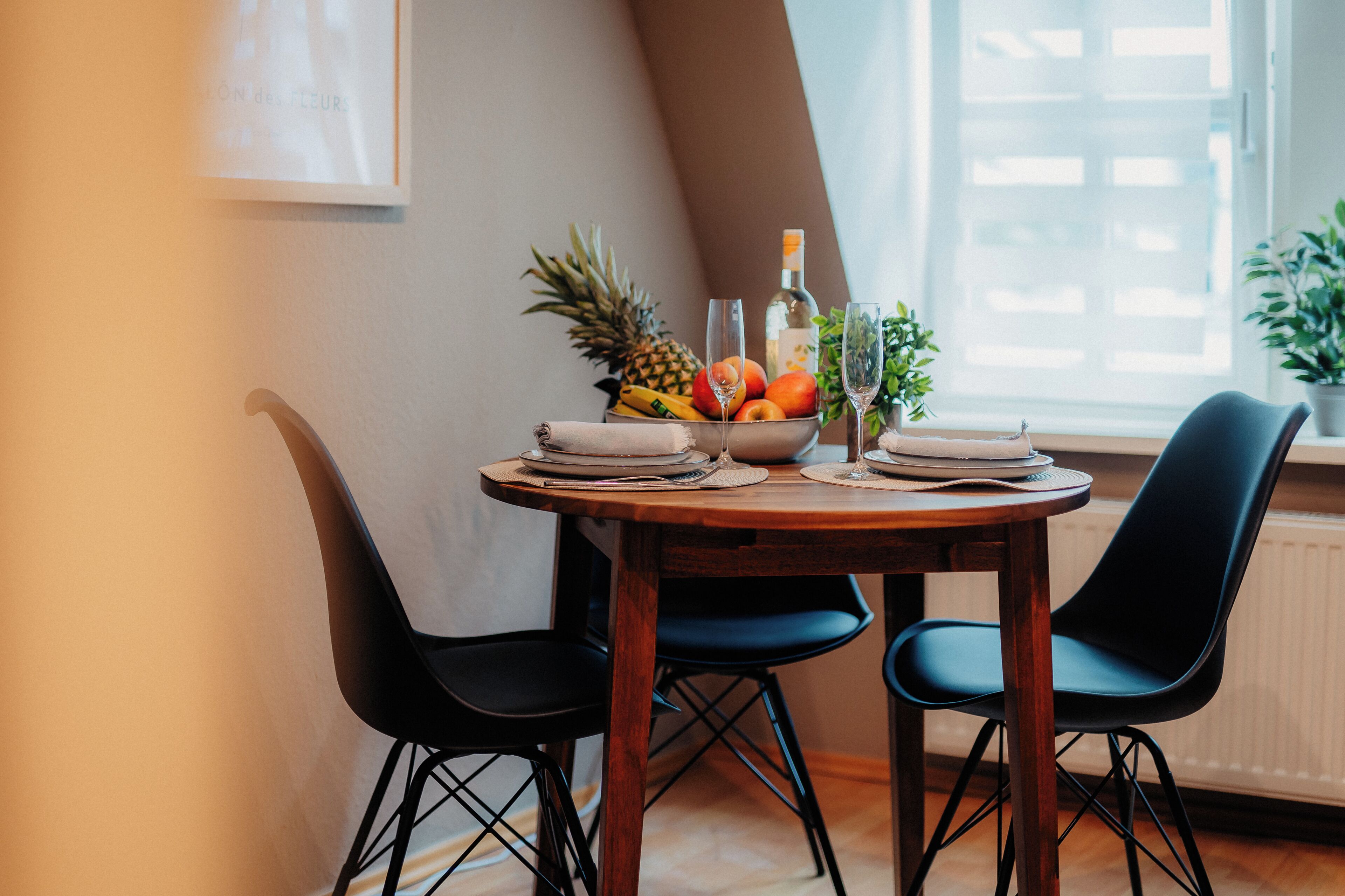 A dining table with a fruit bowl, wine glasses, and plates, surrounded by two black chairs in a room with a window and a radiator.