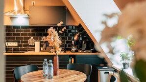 A modern kitchen with a wooden dining table, two chairs, a vase with flowers, and a coffee machine. - Luga Homes - Chausseehaus (Leipzig)