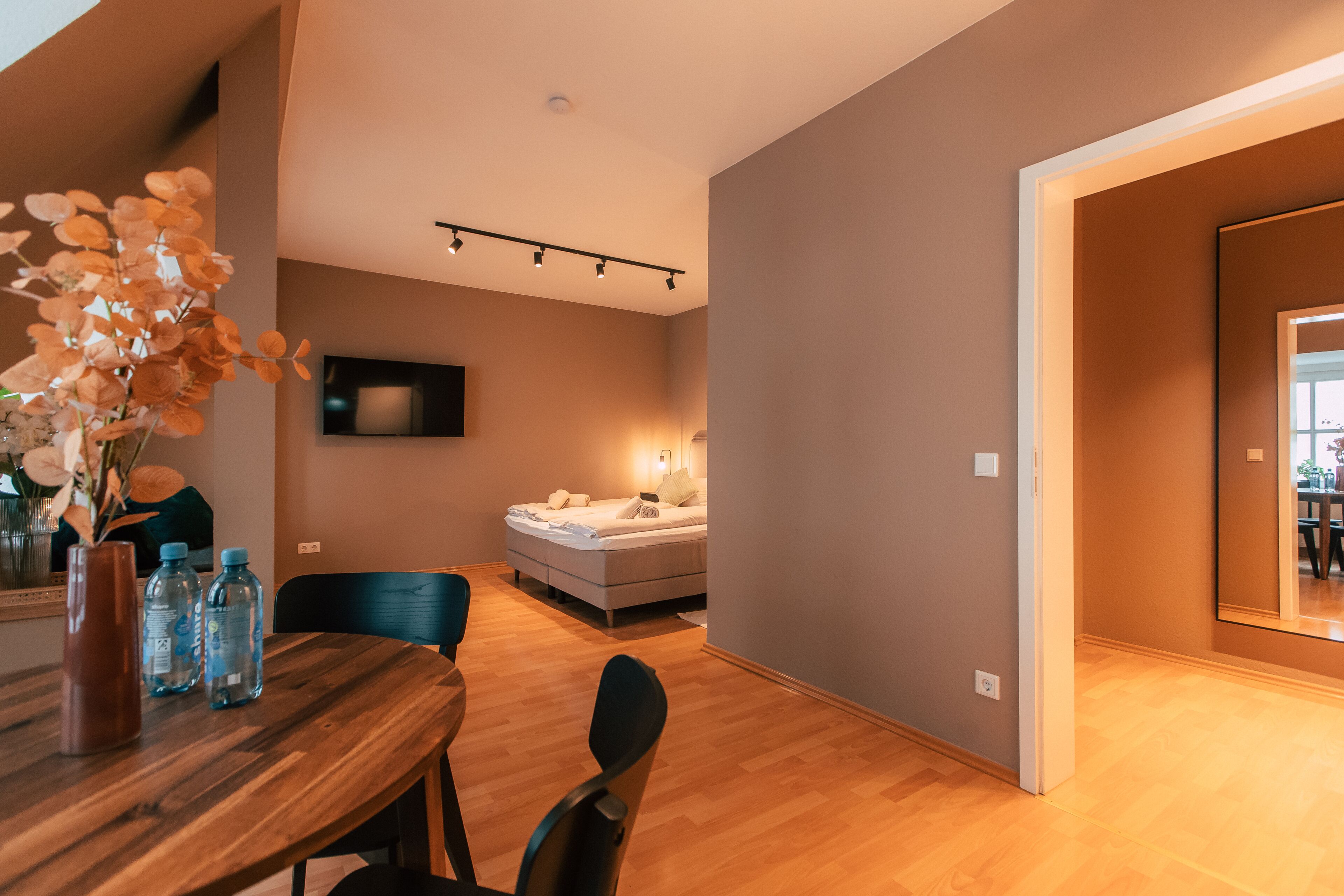 Una habitación de hotel moderna con una mesa de comedor de madera, sillas, una cama y un televisor montado en la pared.