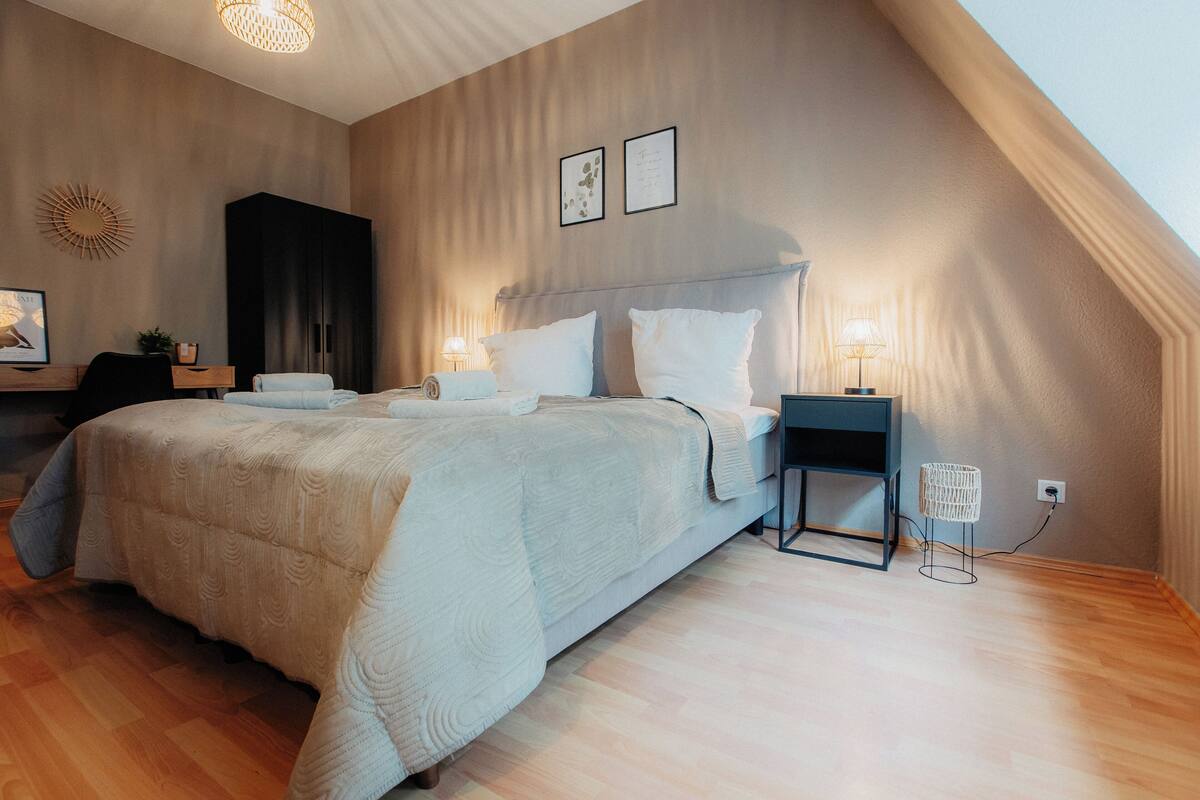 A bedroom with a large bed, a desk, and a chair. There are two bedside tables with lamps.