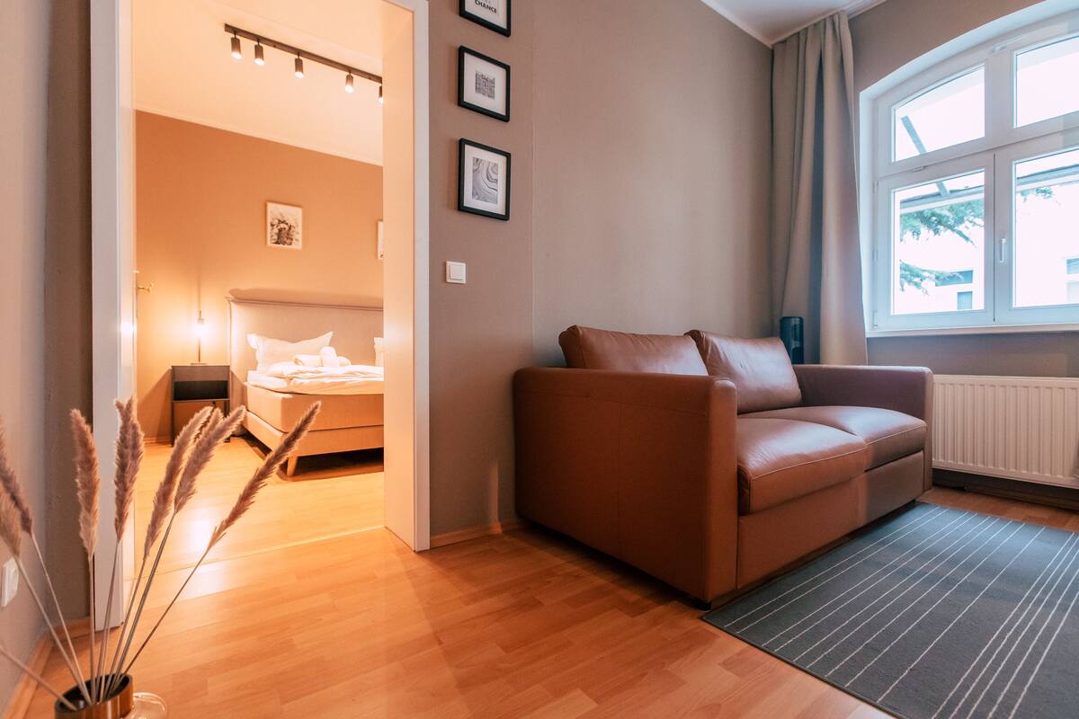 A modern living room with a leather sofa, a wooden floor, a window with curtains, and a bed in the background.
