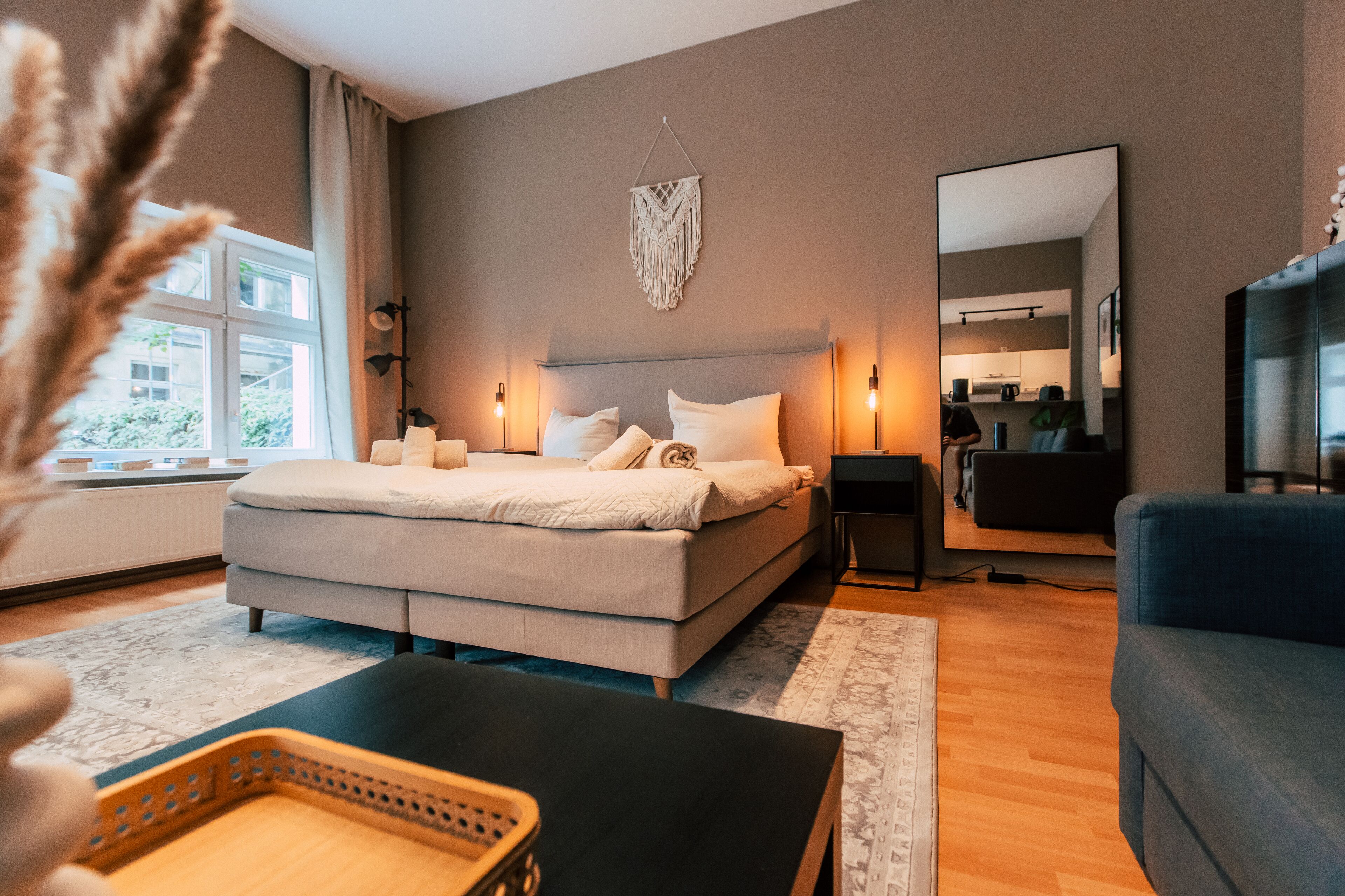 A modern bedroom with a large bed, bedside lamps, a mirror, and a view of the kitchen.