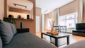 A modern living room with a sofa, a coffee table, a TV, and a dining area with chairs and a table. - Luga Homes - Chausseehaus (Leipzig)