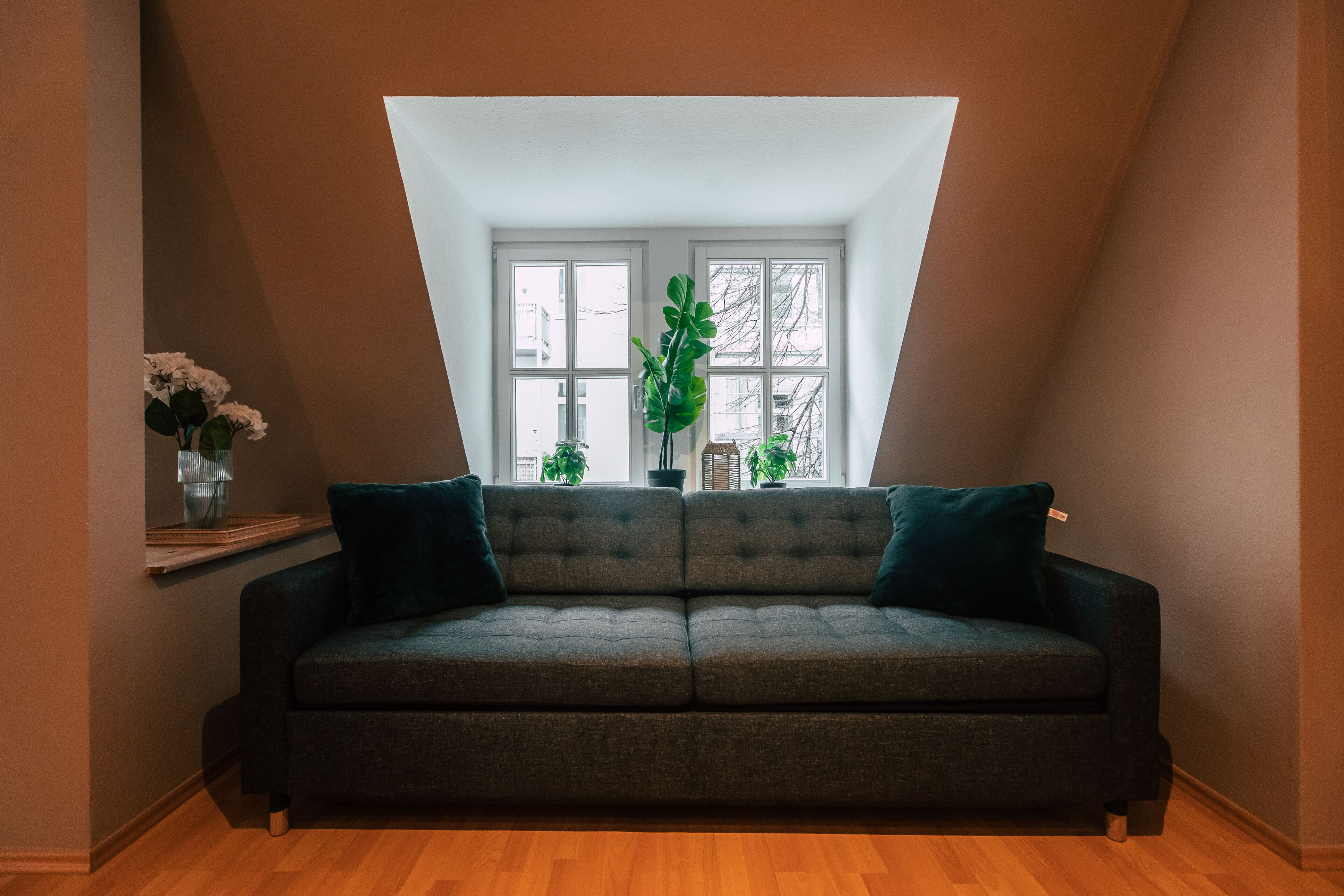 Una habitación con un sofá verde oscuro, una mesita con un jarrón de flores y un ventanal con plantas.