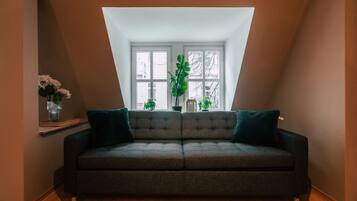 Una habitación con un sofá verde oscuro, una mesita con un jarrón de flores y un ventanal con plantas.