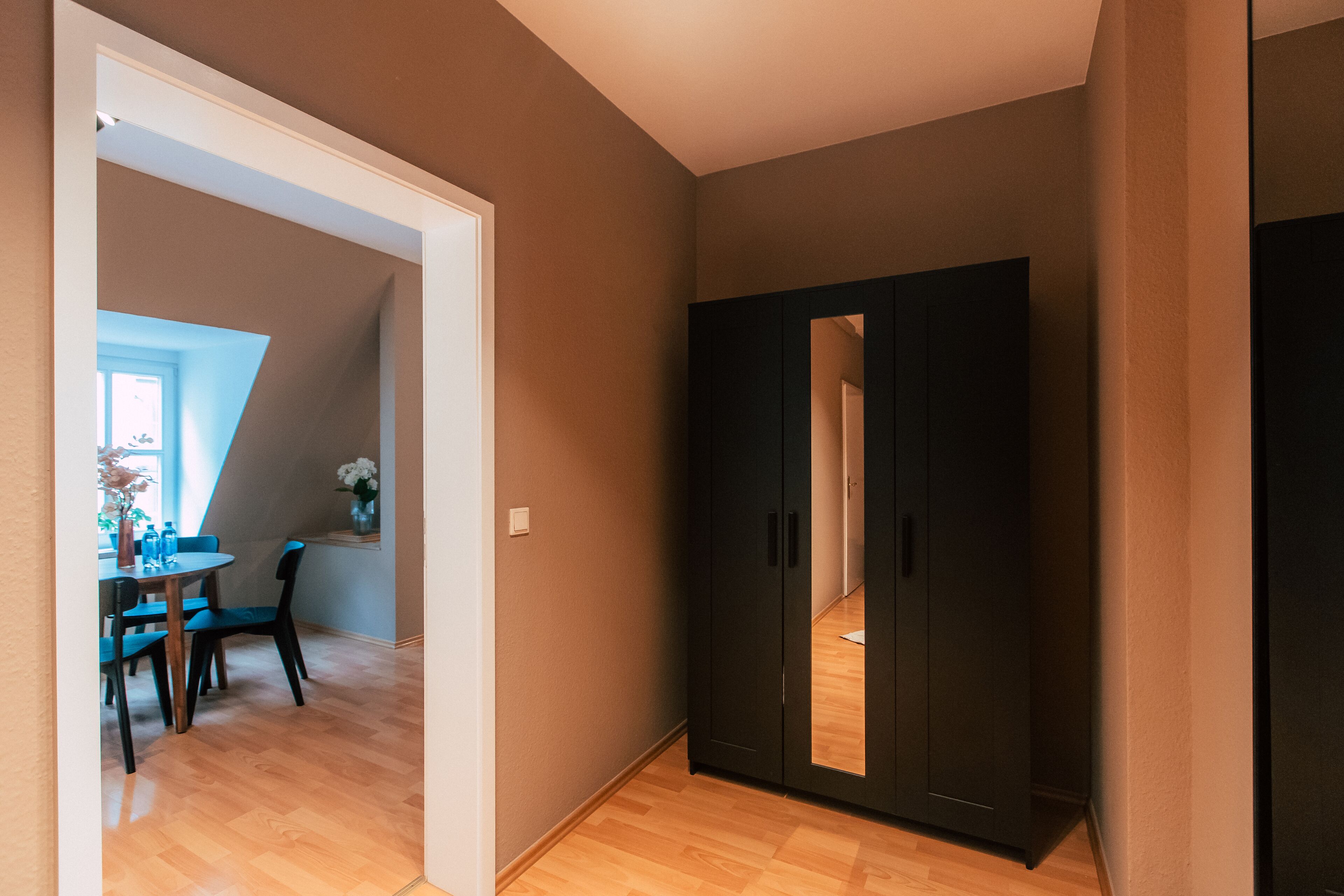 A hallway with a wooden floor, a black wardrobe, and a dining area with a table and chairs.