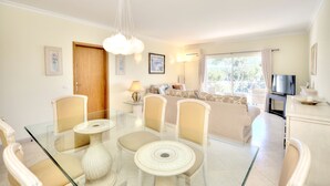 Una habitación bien iluminada con una mesa de vidrio, sillas de mimbre, un sofá, un televisor y un ventanal con cortinas.